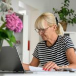 Old woman wearing glasses working on laptop online, sitting at desk on living room