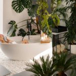 Relaxed young woman bathing in modern bathroom interior decorated with tropical plants